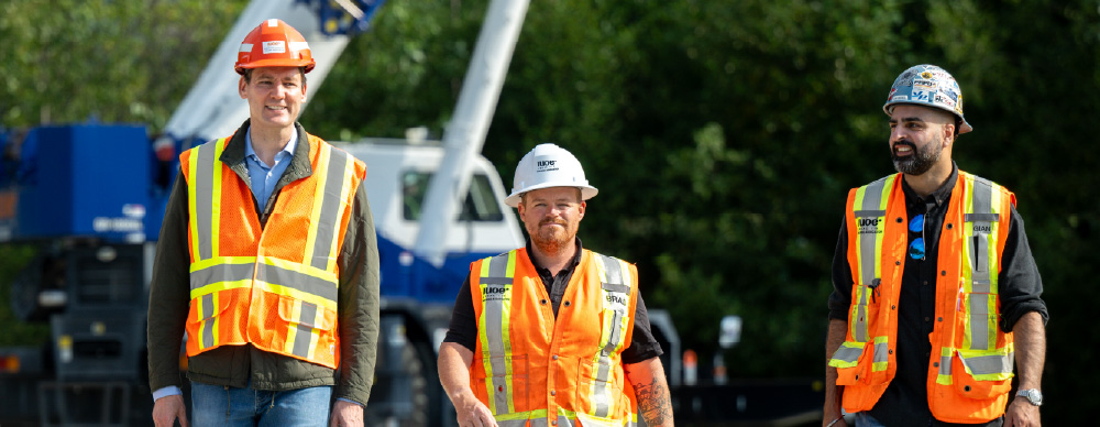 Premier David Eby and two workers in high-viz vests and hard hats walk on a construction site.
