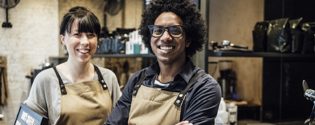 Two smiling baristas stand behind the counter at their coffeeshop.