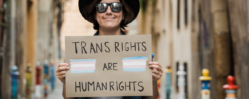 A smiling man in sunglasses holds a homemade sign reading 'trans rights are human rights'